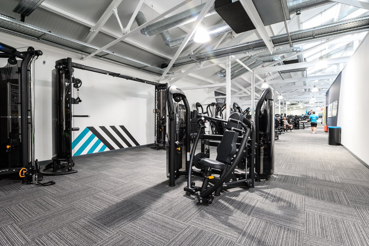 PureGym London East Sheen: Weight machines, resistance equipment, floor mats in a gym facility with industrial ceiling.