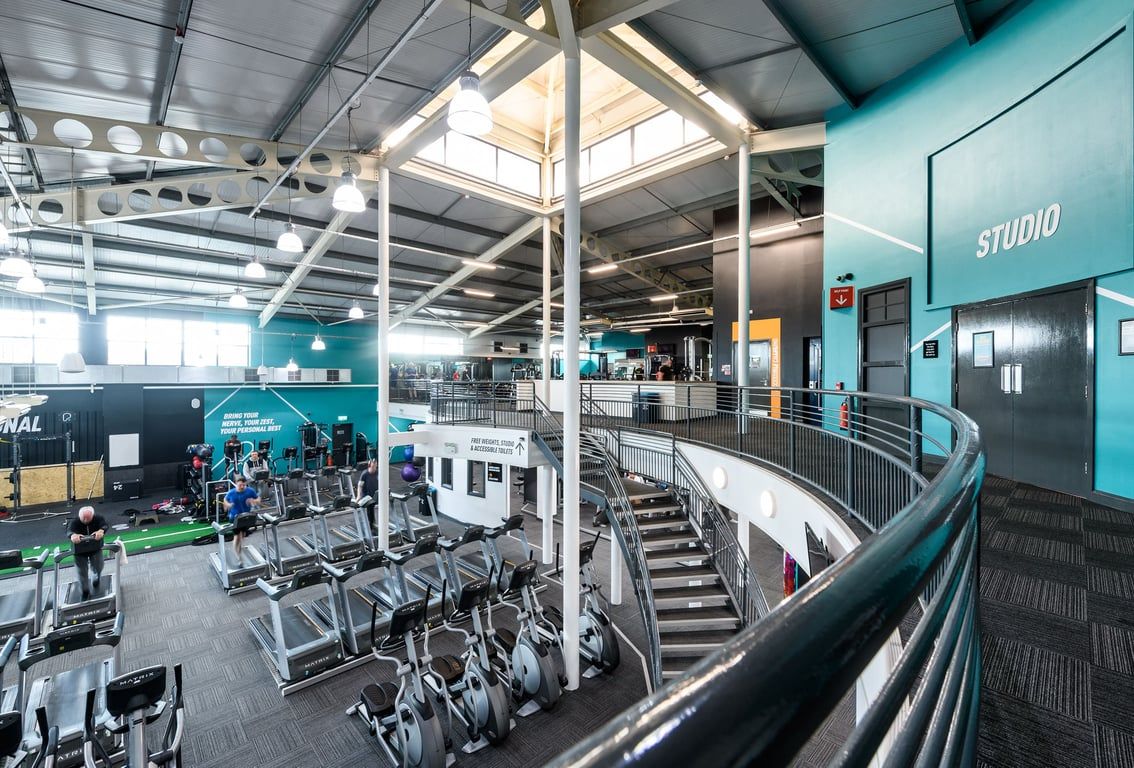 PureGym Manchester Moston: Rows of cardio machines, weight racks, benches, and stairs inside a gym studio.