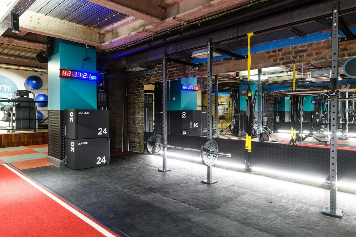 PureGym London Tower Hill: Gym with workout rig, weight racks, rubberized flooring, electronic screens displaying numbers.