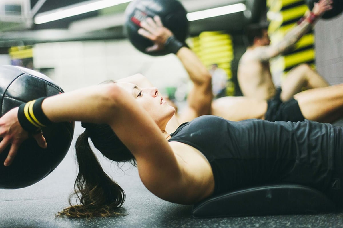 Woman exercising with a medicine ball lying on a mat in a gym with others in the backgroun