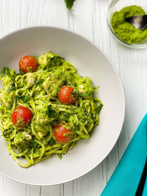 Pasta with green pesto sauce, cherry tomatoes, and fresh herbs in a white bowl.