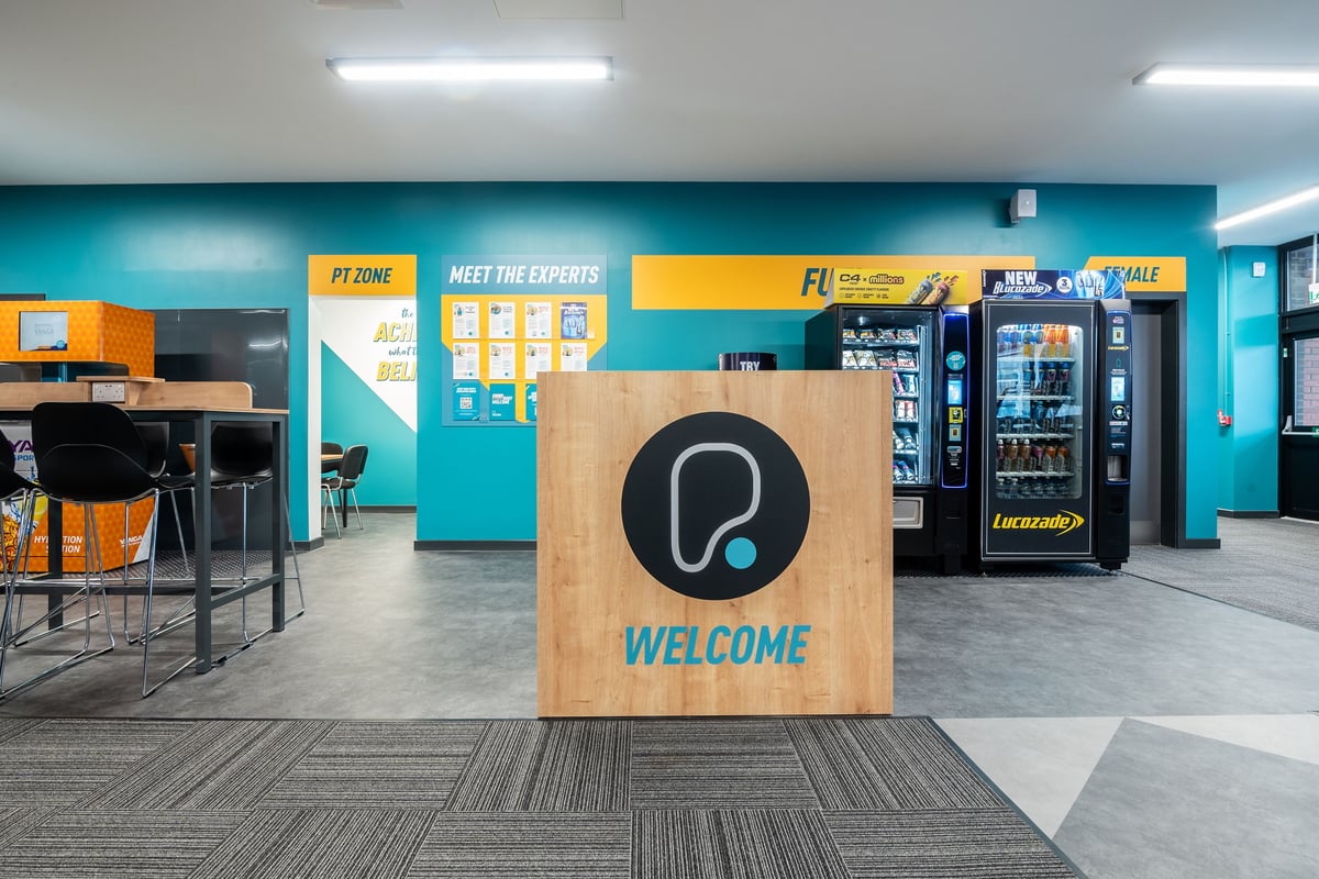 PureGym Airdrie: Vending machines, welcome sign, seating area with bright teal walls.