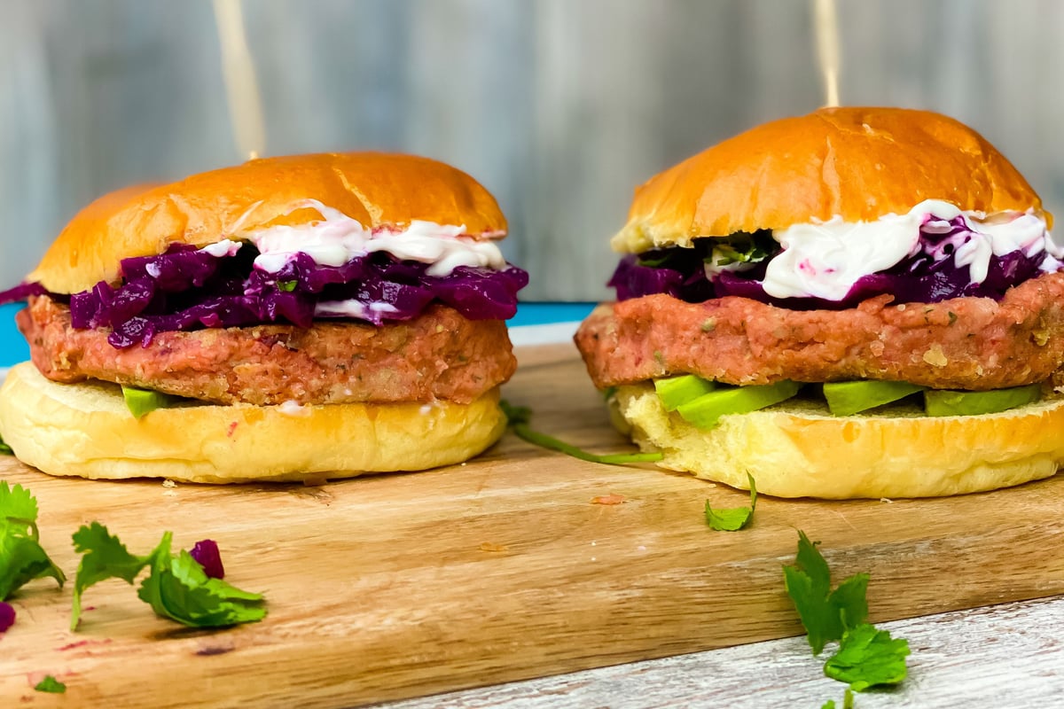 Two vegan burgers with breaded patties, purple cabbage and white sauce on wooden board