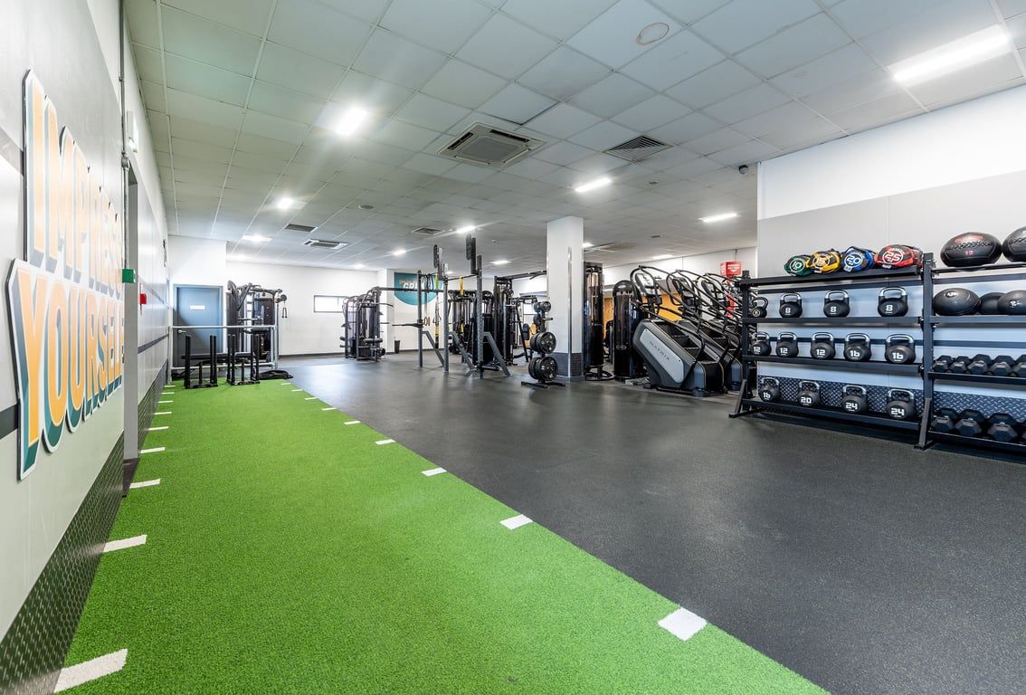 PureGym Northolt: Gym studio with weightlifting racks, free weights, turf area, mirrors, and rubberized flooring.