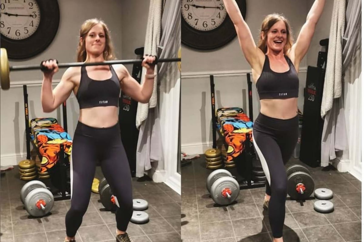 Woman lifting a barbell from shoulder height to overhead in a home gym with weights.