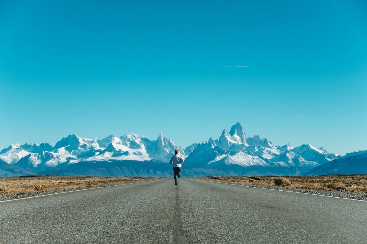 Person running on empty road towards snow-capped mountain range under clear blue sky
