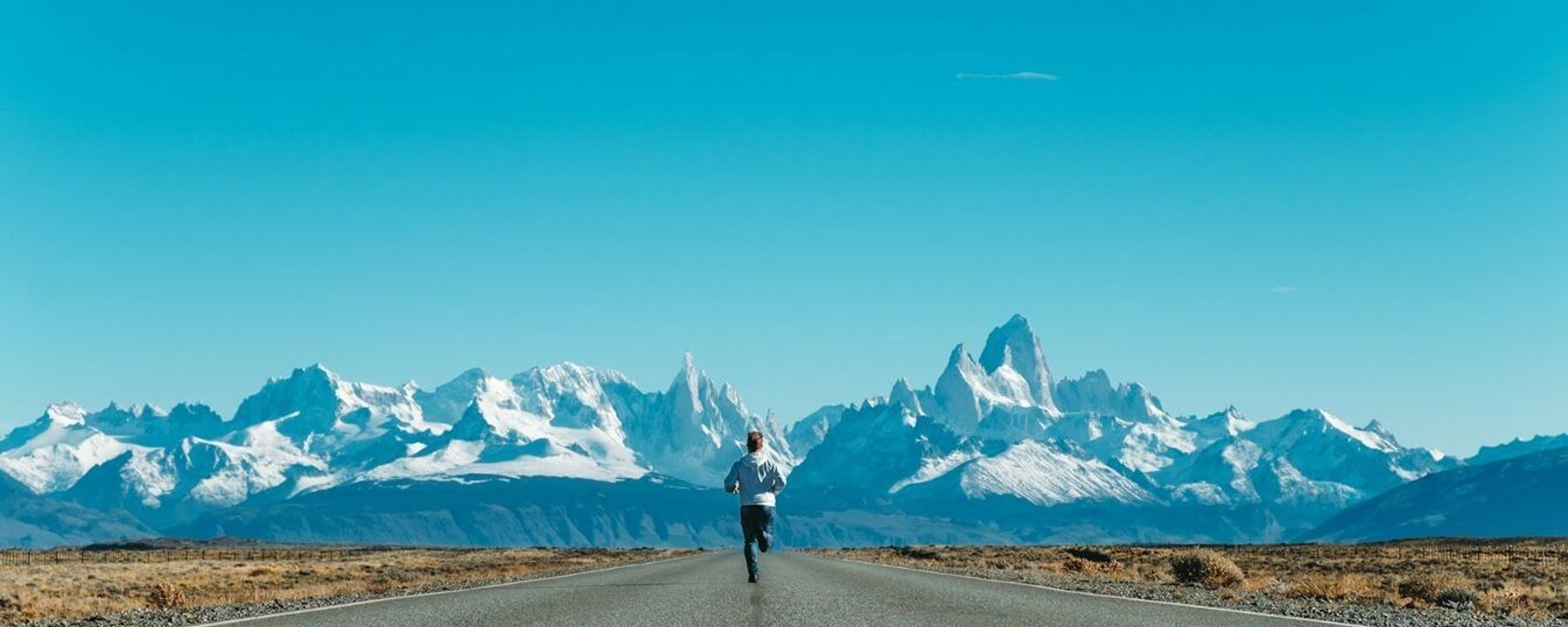 Person running on empty road towards snow-capped mountain range under clear blue sky