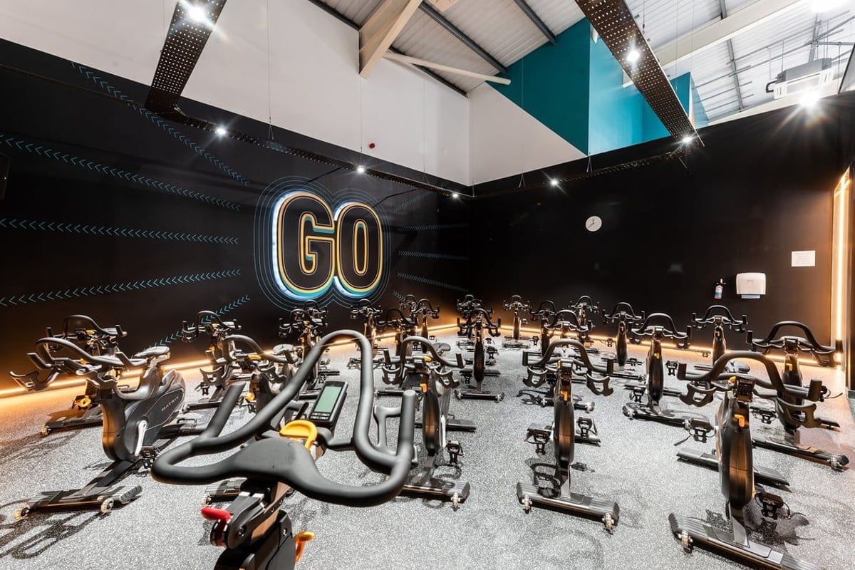 PureGym Macclesfield Silk Road: Rows of stationary bikes, illuminated "GO" signage, dark walls with teal accents.