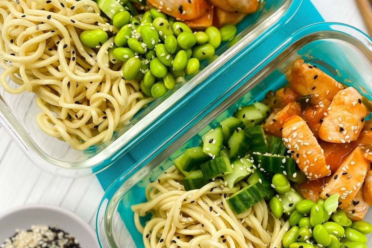 Two glass containers with noodles, diced cucumber, edamame, and sesame chicken pieces
