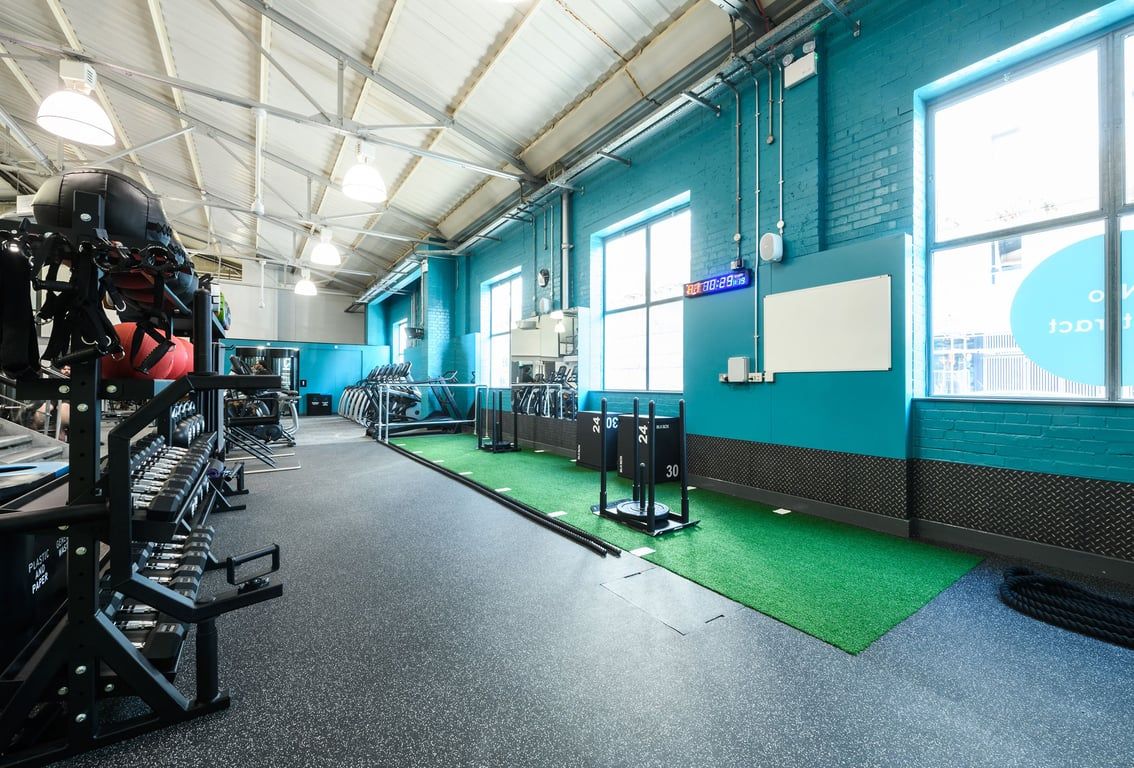 PureGym London Clapham: Teal-walled gym studio with turf area, weightlifting rigs, free weights, and resistance machines.