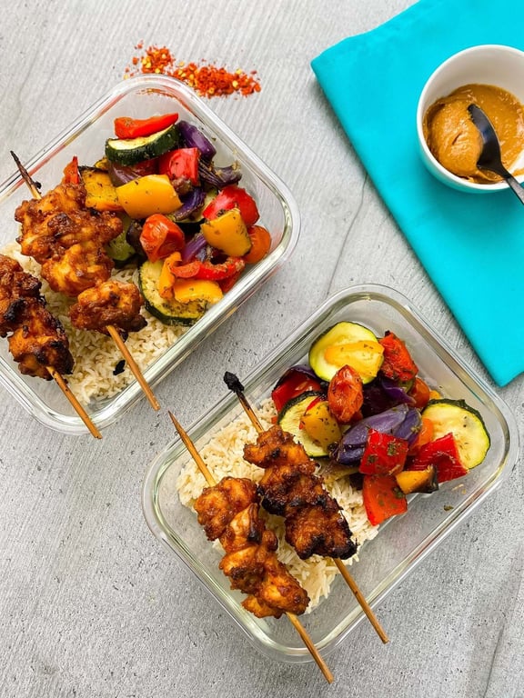 Two glass containers with grilled chicken skewers, rice, and mixed roasted vegetables.