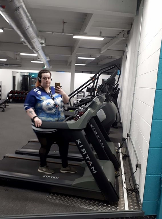Person wearing headphones standing on a treadmill in a gym taking a mirror selfie