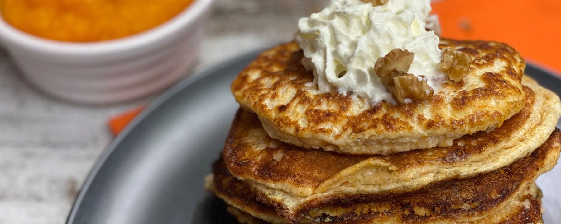 Stack of golden pancakes topped with cream and walnuts beside a bowl of pumpkin purée
