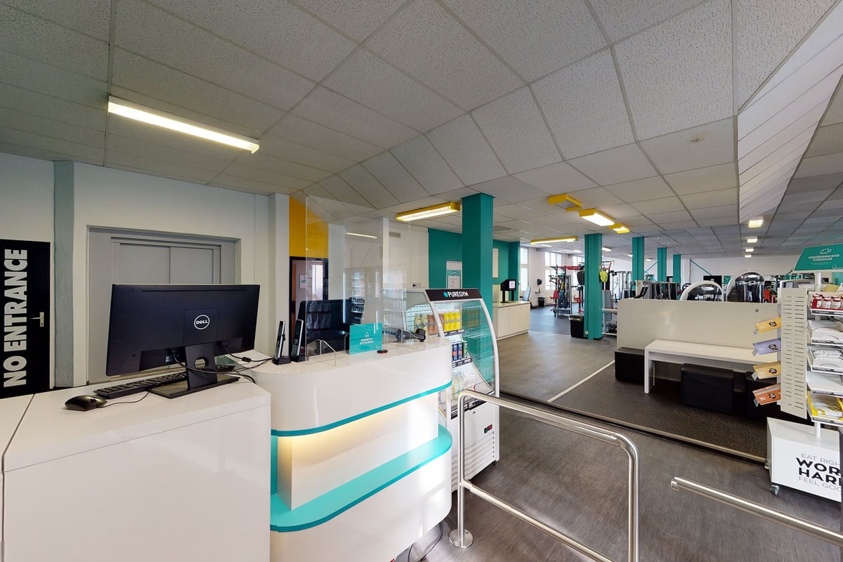 PureGym Rorschach: Zone de réception avec bureaux, ordinateurs et signalisation colorée indiquant les différentes zones de fitness ou studios.