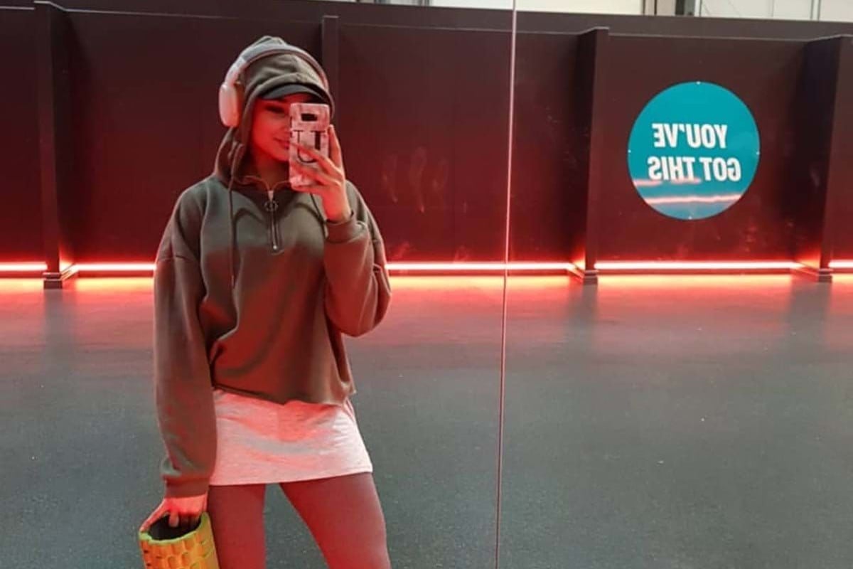 Woman in sportswear holding a foam roller taking a mirror selfie in a gym room