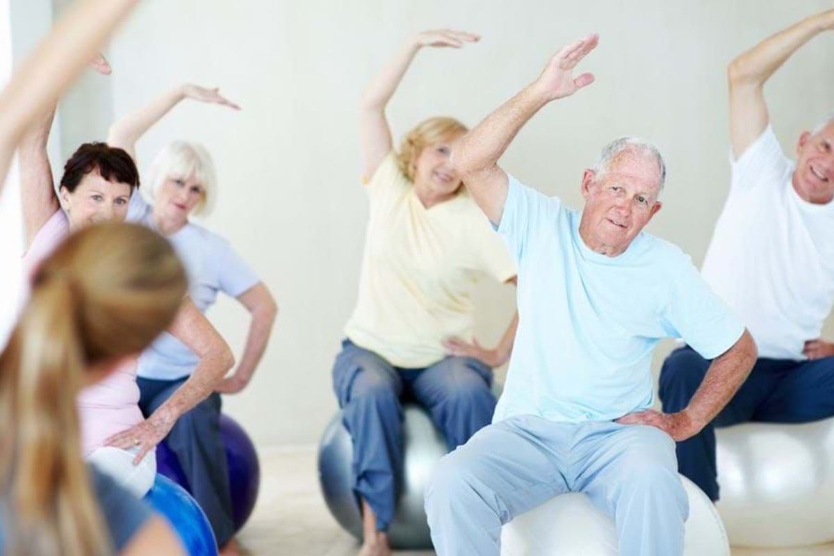 Seniors sitting on exercise balls stretching arms sideways in a fitness class.
