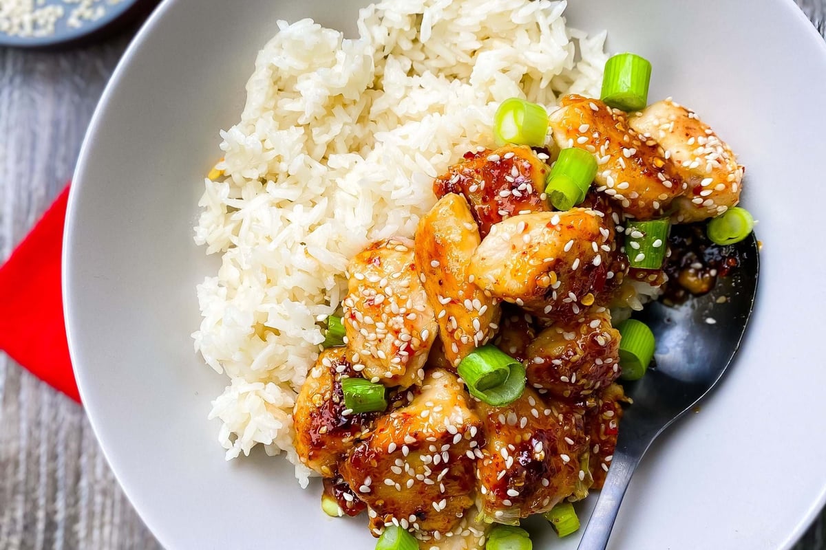 White rice with sesame chicken and chopped spring onions in a bowl with a spoon
