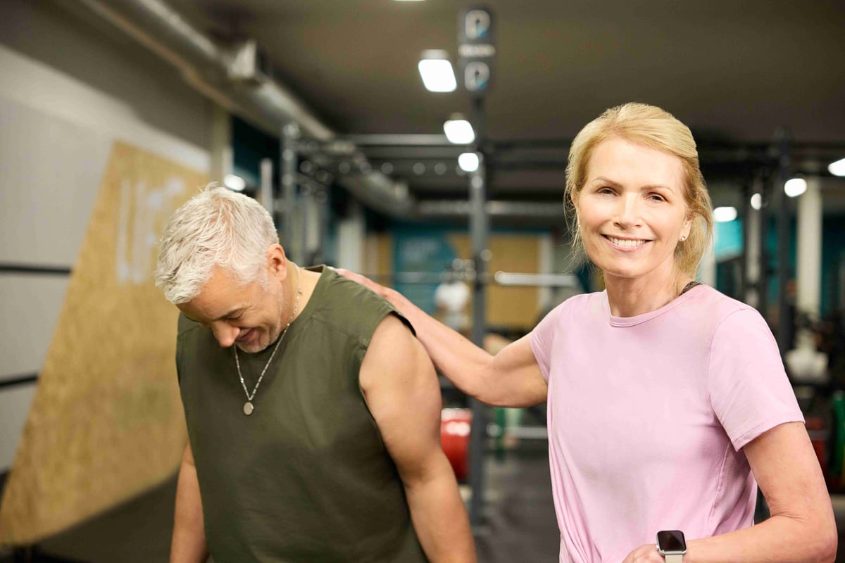 Smiling woman in pink shirt with hand on shoulder of man in green sleeveless top in gym