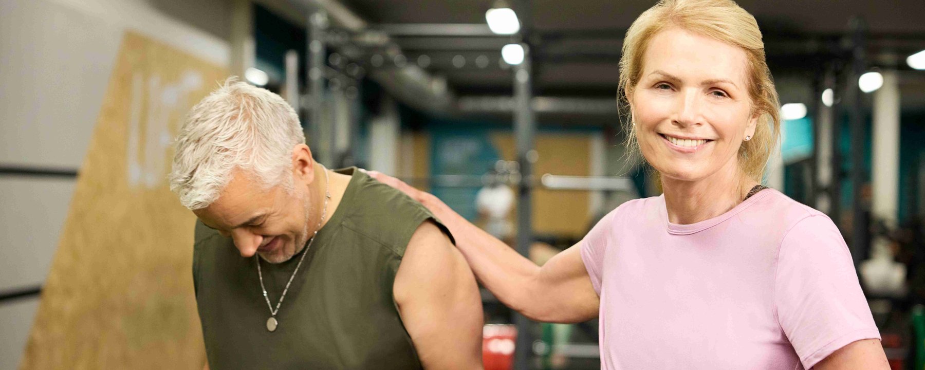 Smiling woman in pink shirt with hand on shoulder of man in green sleeveless top in gym