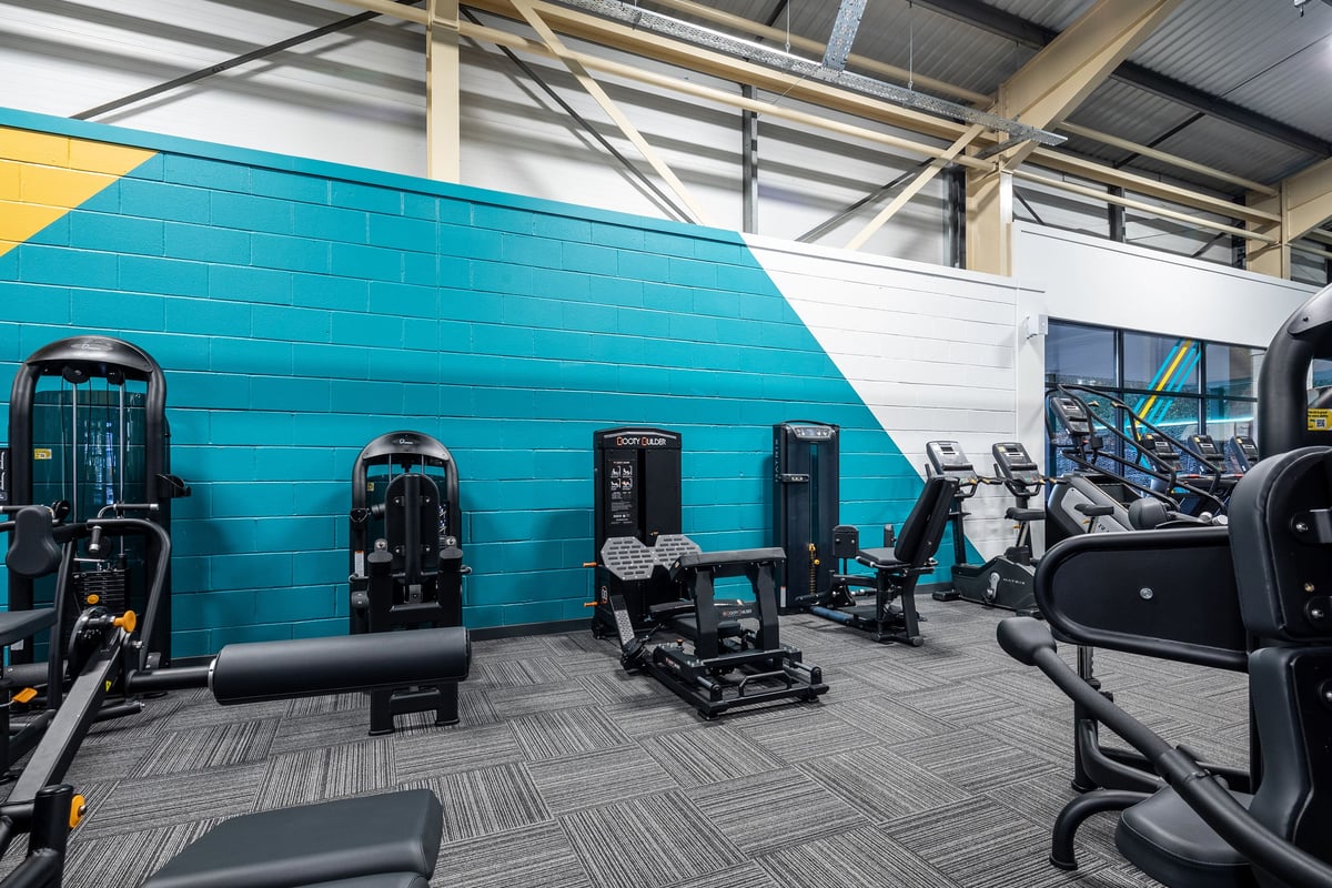 PureGym Neath: Rows of strength machines, cable crossovers, free weights, and exercise benches in a teal and white gym.