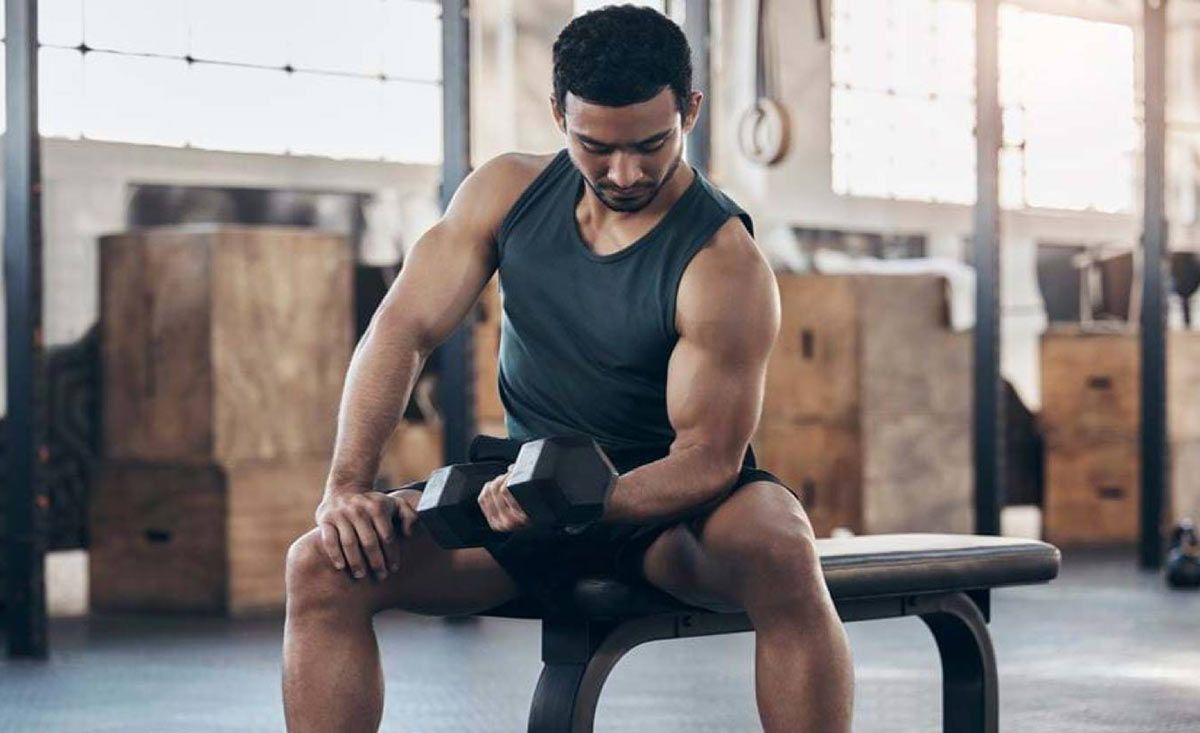 Mann im schwarzen Tanktop hebt Kurzhantel auf Fitnessbank sitzend