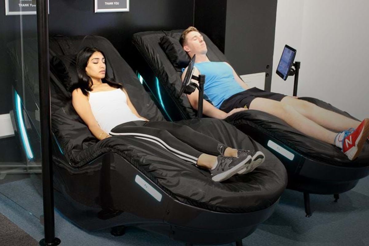 Two people relaxing on black massage chairs with screens attached in a room
