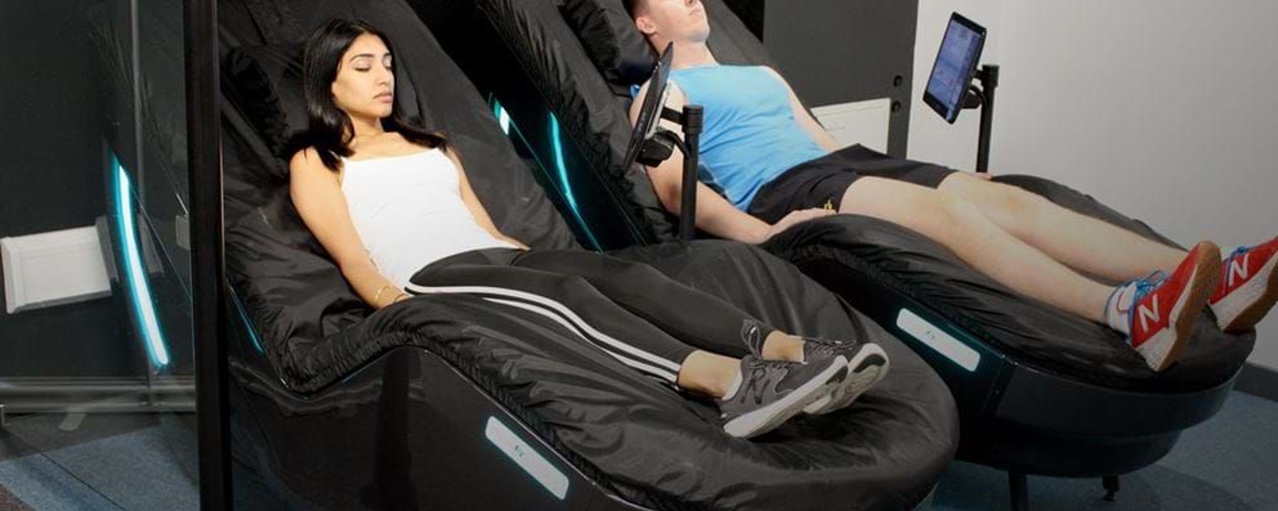 Two people relaxing on black massage chairs with screens attached in a room
