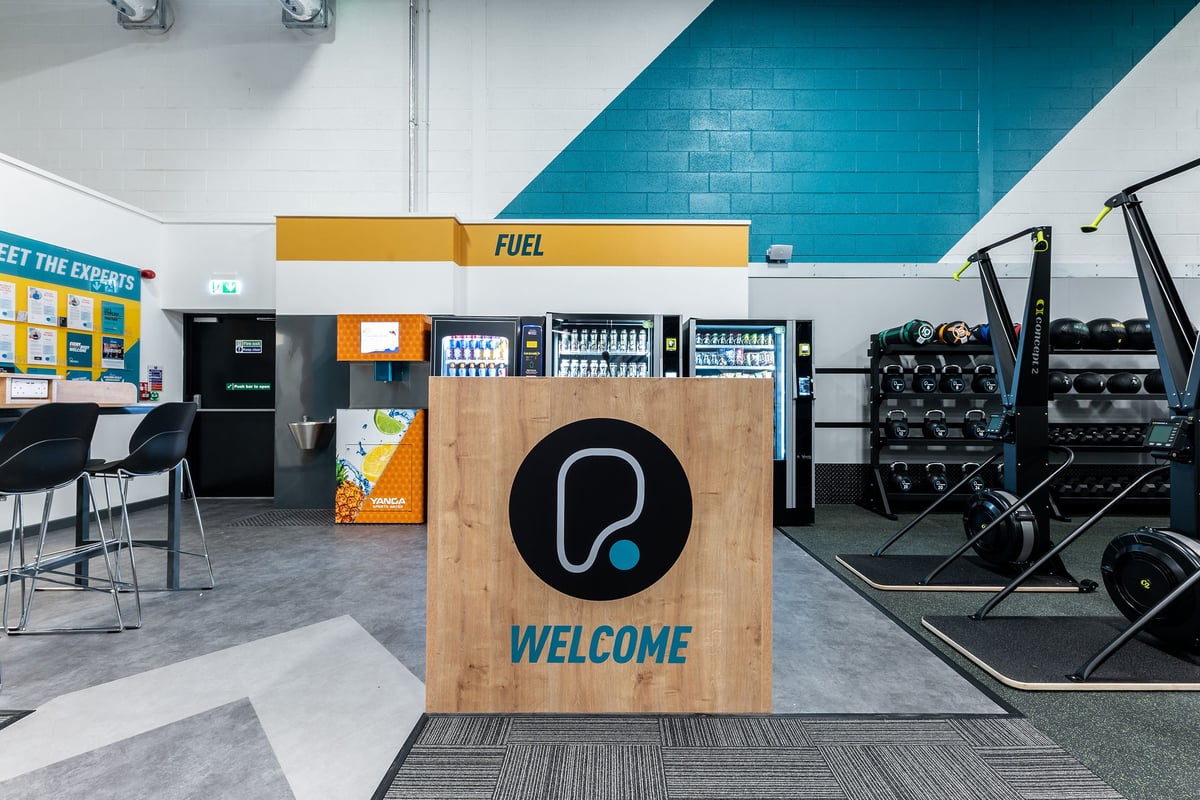 PureGym Ballymena: Free weights, resistance machines, vending area with teal tiled wall and wood welcome sign.
