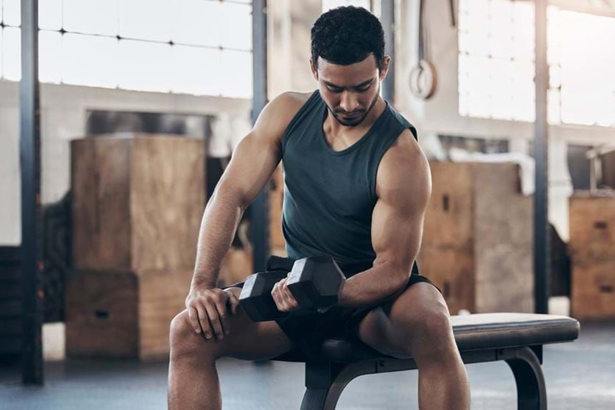 Man in black tank top lifting dumbbell while sitting on gym bench