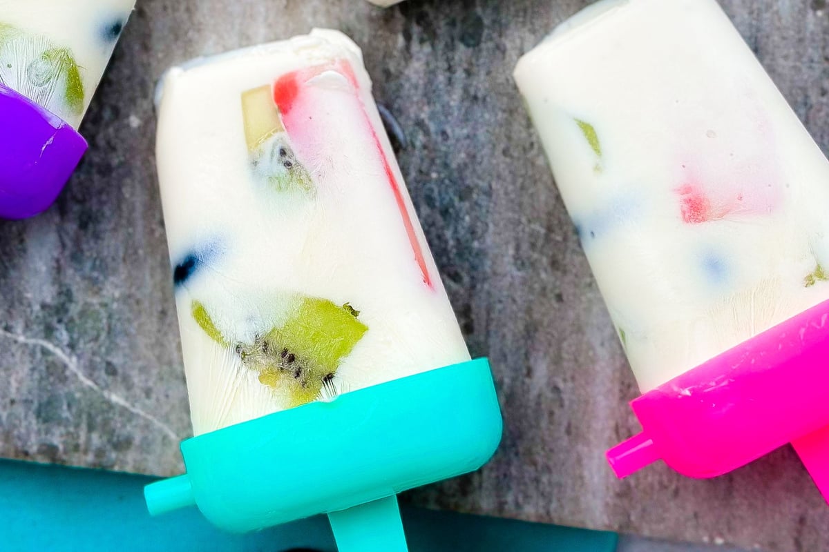 Two frozen yoghurt lollies with colourful fruit pieces on a grey marble surface