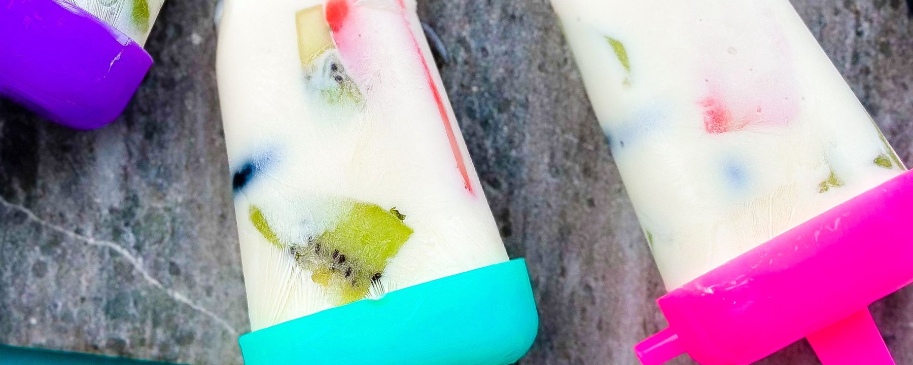 Two frozen yoghurt lollies with colourful fruit pieces on a grey marble surface