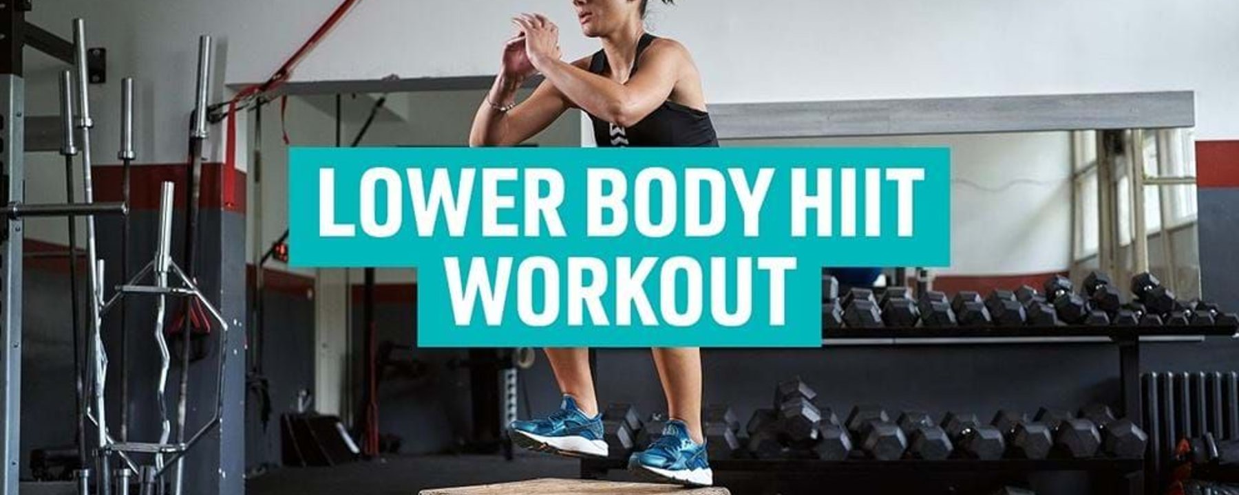 Woman jumping on wooden box in gym with dumbbells and barbells in background