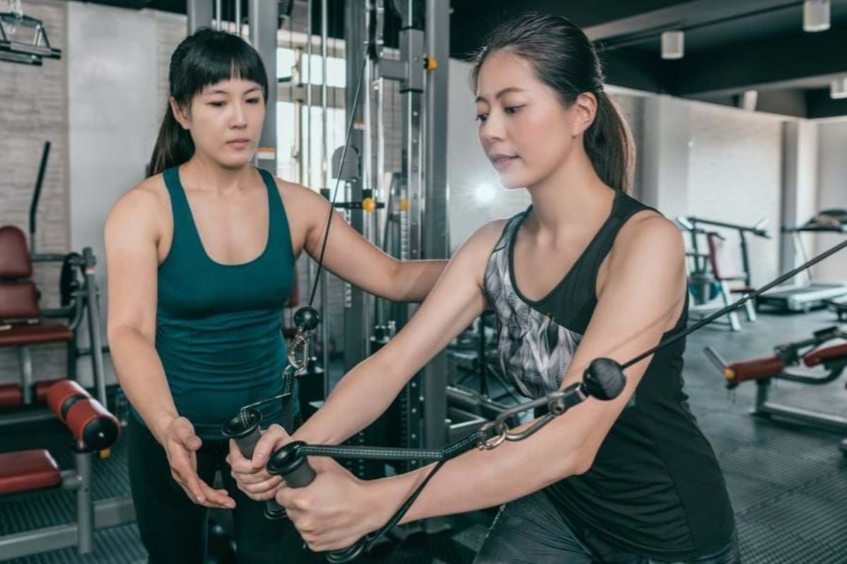 Woman training on cable machine with trainer assisting in gym.