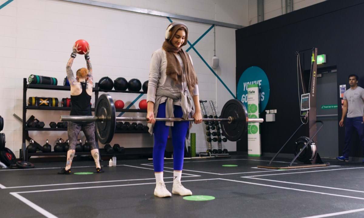 Woman in hijab lifting barbell while another person holds a red medicine ball in gym