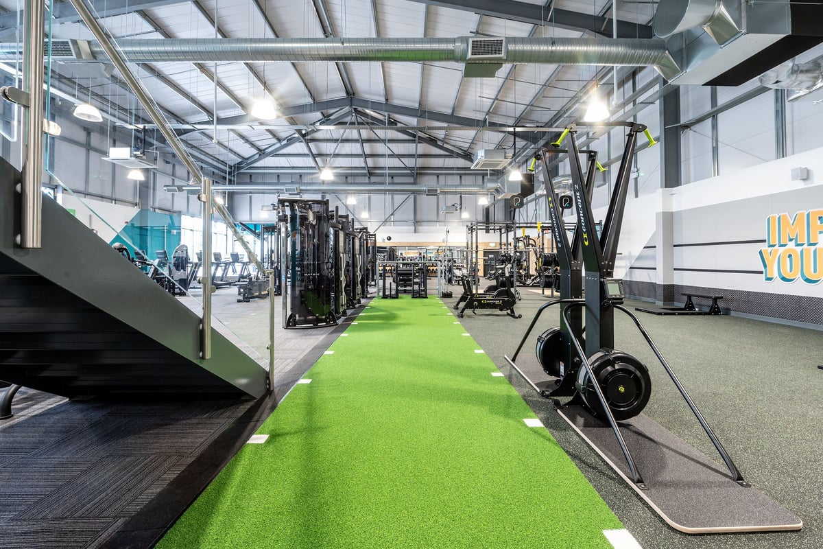 PureGym Cirencester: Strength racks, barbells, weight plates, green turf area, and mirrored walls in a gym studio.