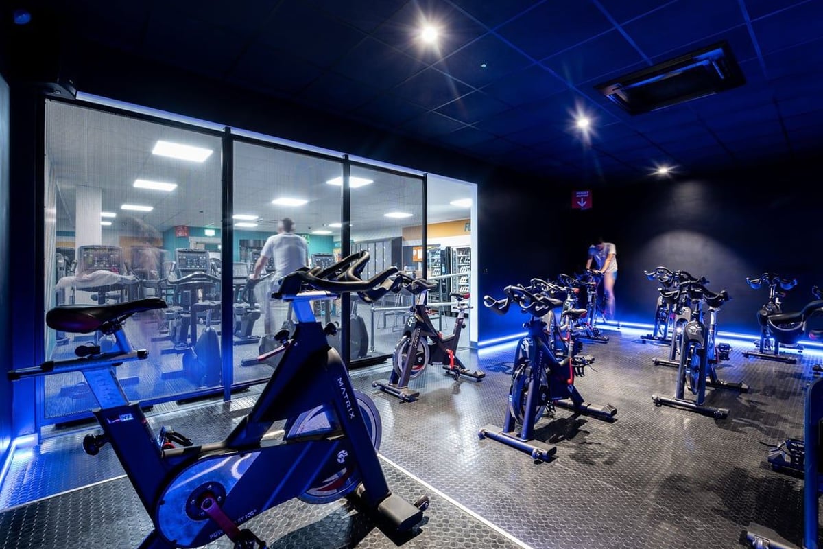 PureGym Epsom: Row of exercise bikes, treadmills behind glass wall, dim lighting with blue accents.