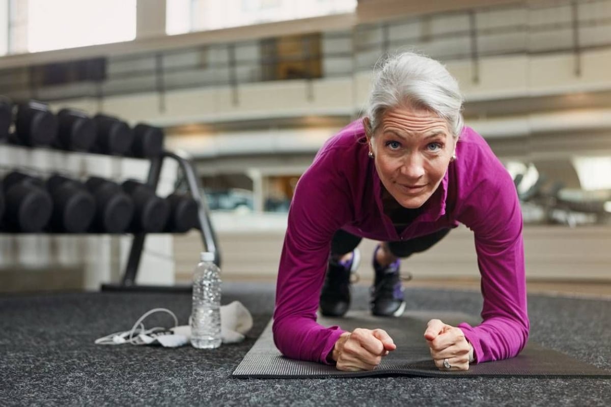 Ältere Frau in violetter Jacke macht eine Plankenübung auf einer Matte im Fitnessstudio