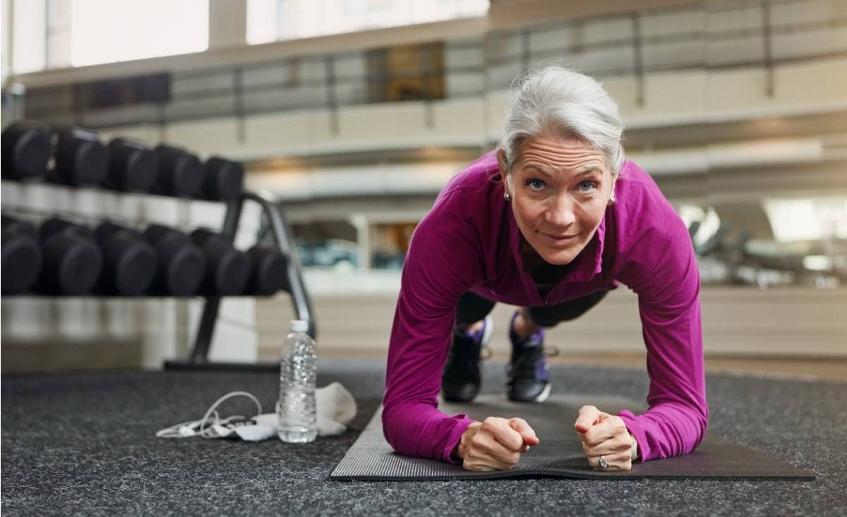Ältere Frau in lila Oberteil macht Unterarmstütz auf Matte im Fitnessstudio mit Gewichten