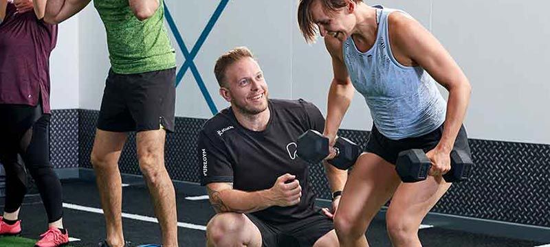 Mann coacht Frau beim Hanteltraining im Fitnessstudio, andere trainieren daneben