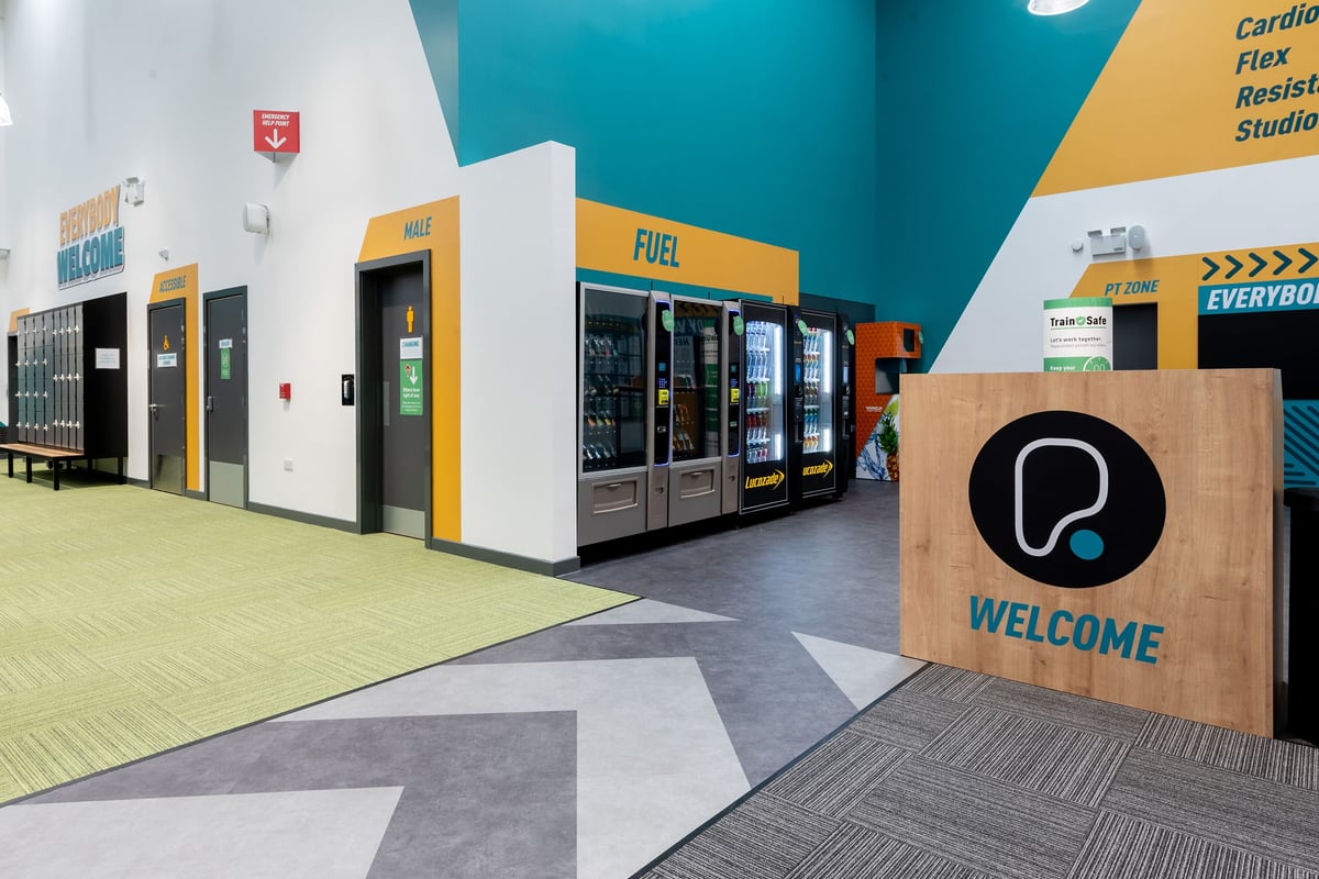 PureGym Colchester Retail Park: Vending machines, welcome sign, open tiled space - looks like an entrance area or lobby.