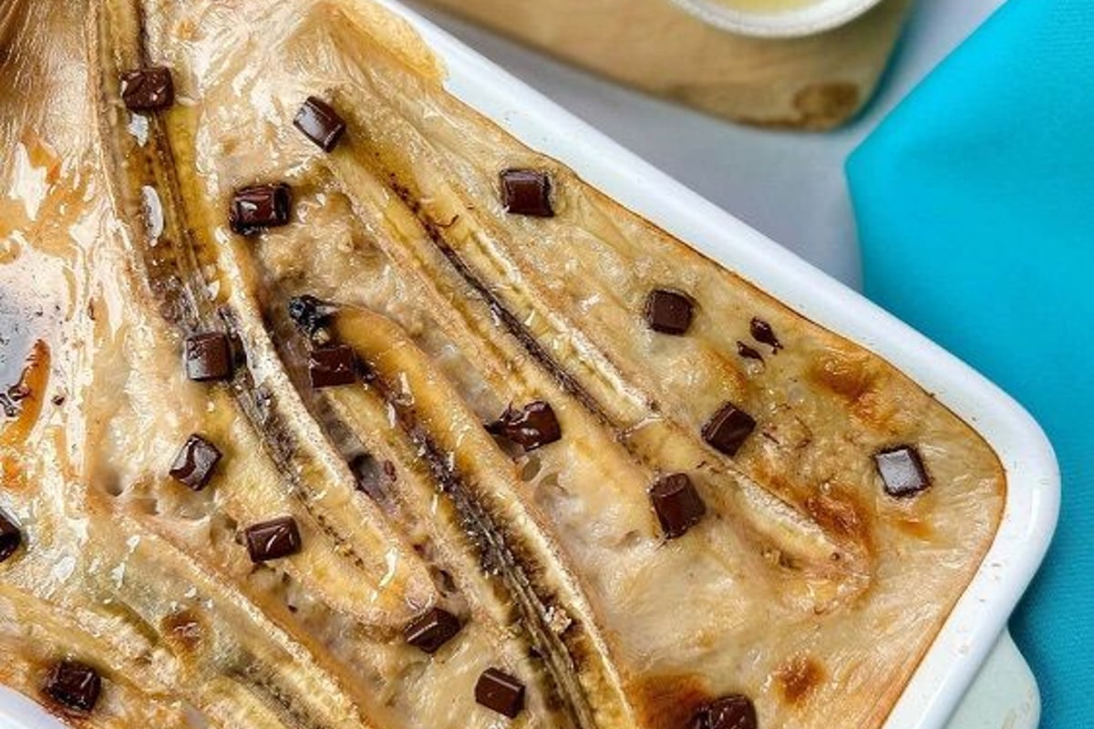 Baked banana dessert with chocolate chunks in a white dish beside honey and cream bowls