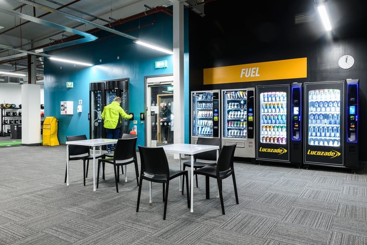 PureGym Cambridge Grafton Centre: Vending machines displaying sports drinks and nutritional products, tables and chairs.