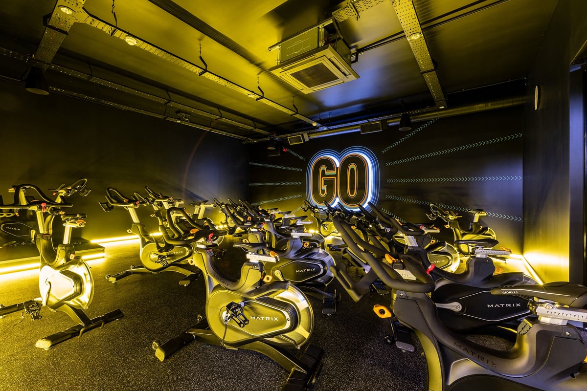 PureGym Doncaster: Rows of stationary bikes in a dimly lit space with neon "GO" sign and exposed ceiling pipes.