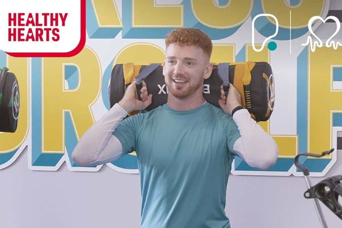 Man lifting weighted sandbag in gym with Healthy Hearts logo and mural behind him