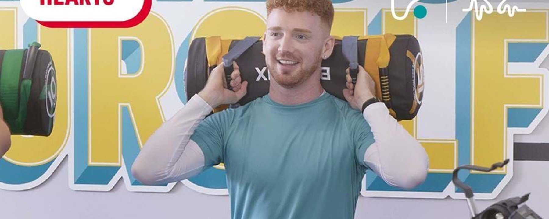 Man lifting weighted sandbag in gym with Healthy Hearts logo and mural behind him
