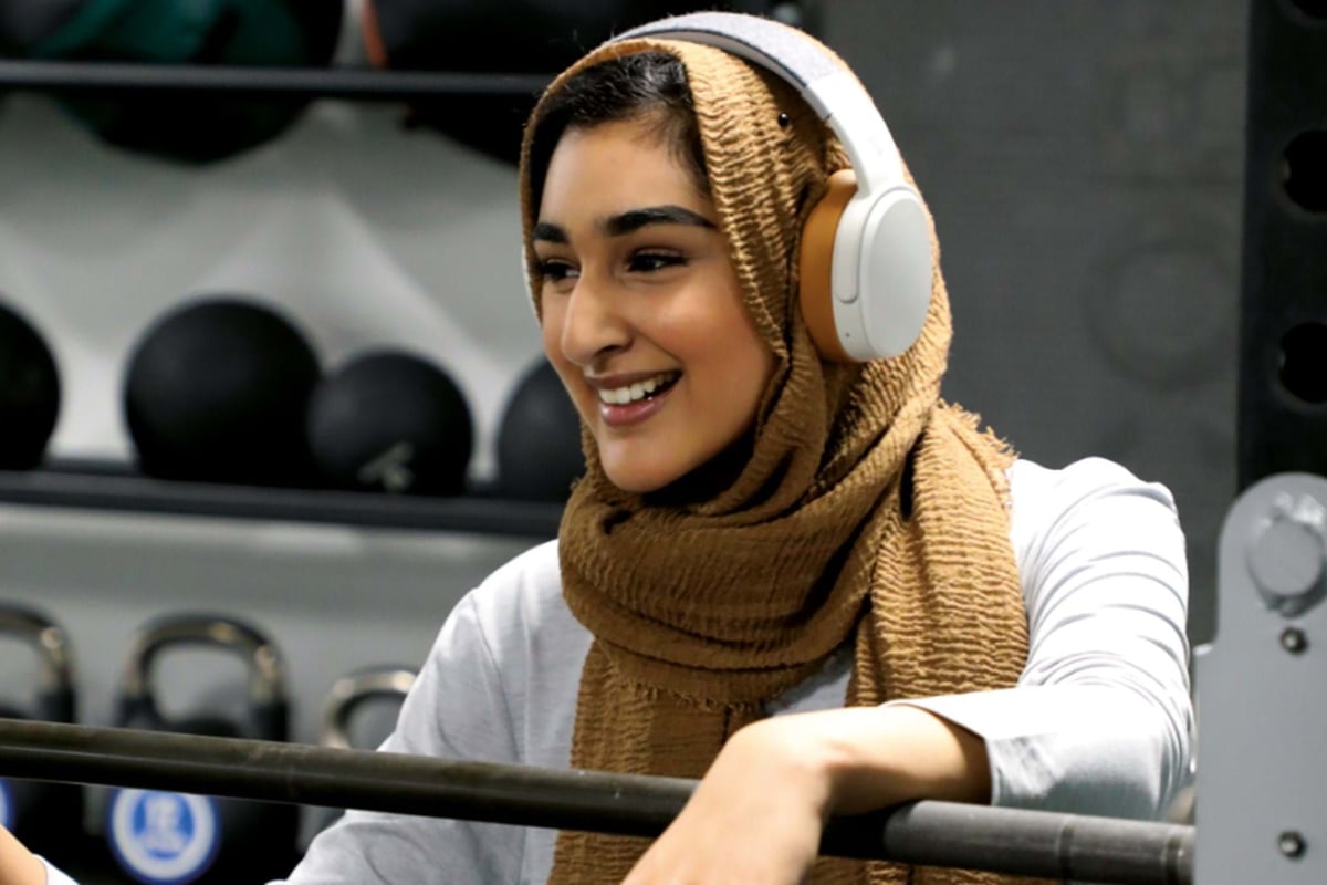 Smiling woman wearing a brown hijab and white headphones holding a barbell in gym