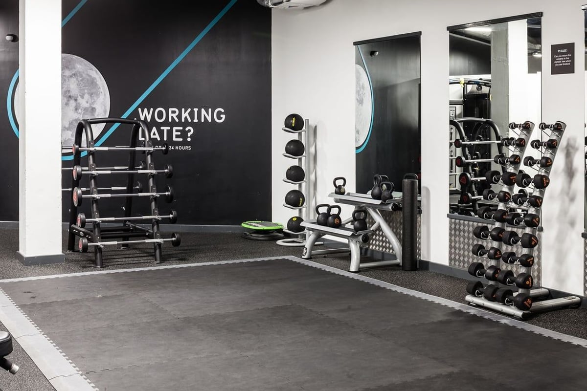 PureGym Manchester Stretford: Weightlifting racks, free weights, benches, and mirrored walls in a dimly lit gym space.