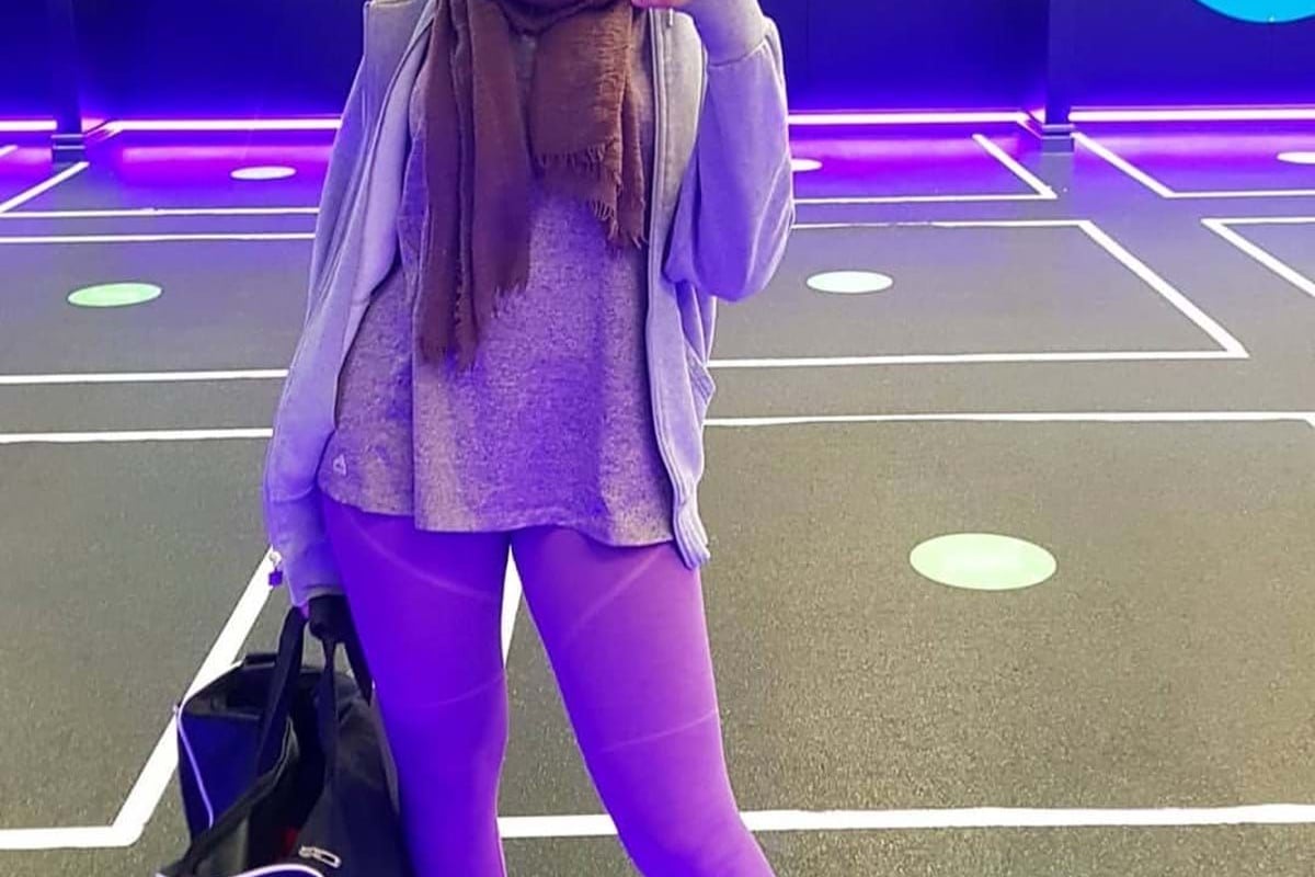 Woman in purple leggings and brown scarf taking a mirror selfie in a gym with neon lights