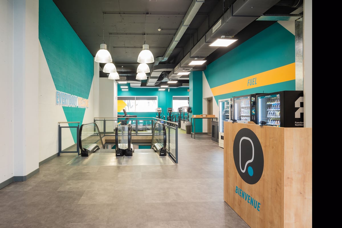 PureGym La Chaux-de-fonds: Cardio machines, wooden kiosks, teal walls, and pendant lighting.
