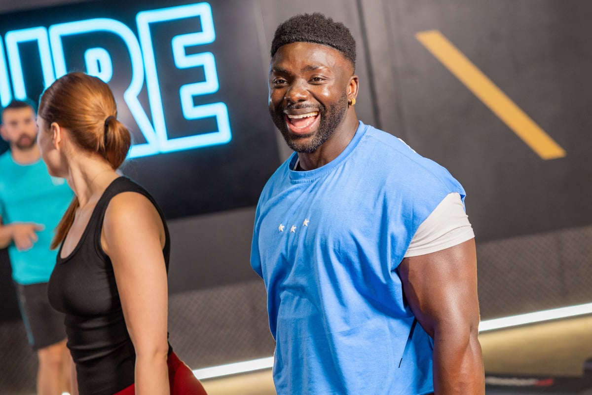 Smiling man in blue sleeveless shirt with two people in the background at gym